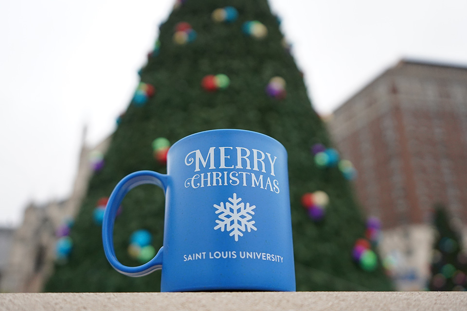 A mug says "Merry Christmas Saint Louis University" with an image of a snowflake on it as it rests in front of a large Christmas tree outside.