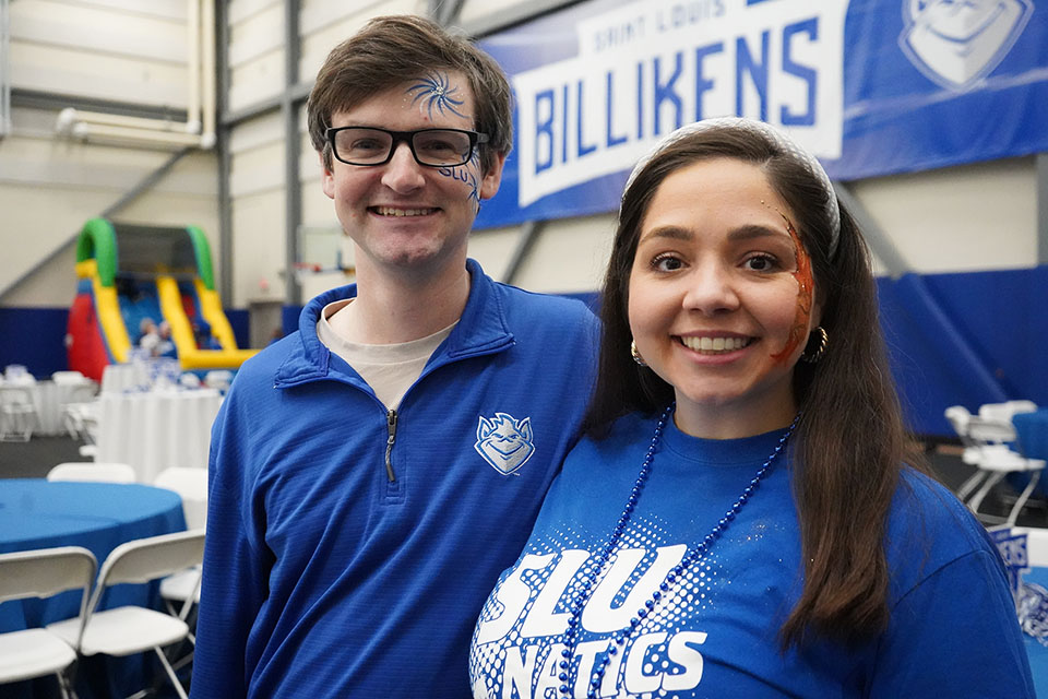 Two people pose for a photo wearing blue with basketball and Billiken face paint on their cheeks.