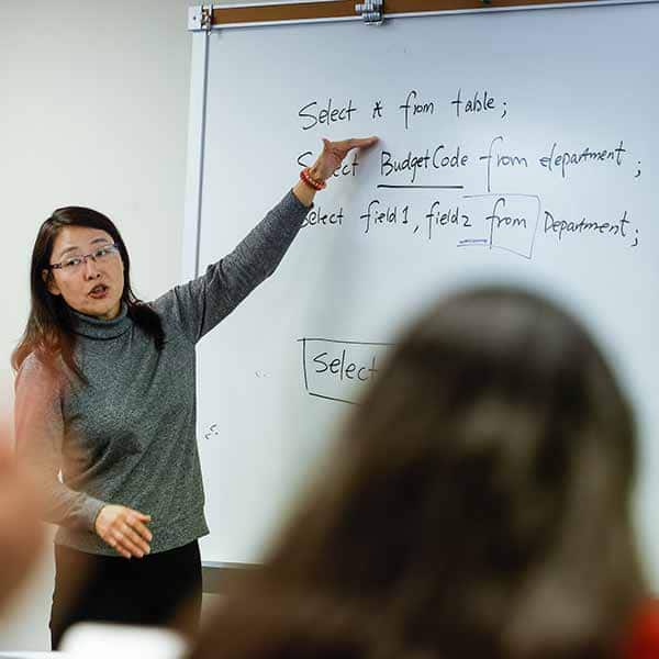 A female faulty member points to instructions on a whiteboard. Heads of students in the classroom can be seen in the foreground.