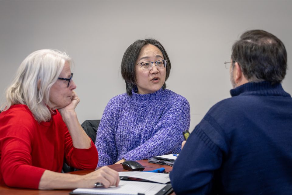 a women in the middle of two other people sits talking at a desk