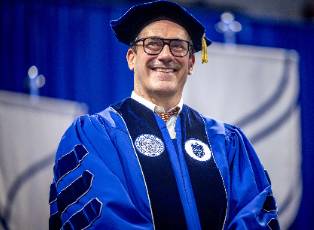 Actor Jon Hamm wearing a graduation cap and gown smiles while standing in front of SLU logos on a stage.