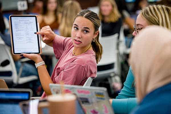 A female student holds up a tablet computer to show her work to two other students.