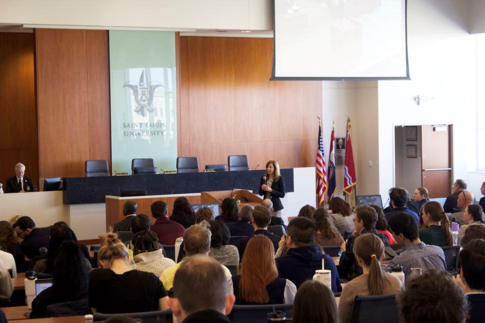 Audience members are seen from behind as a speaker talks at the front of a lecture hall.