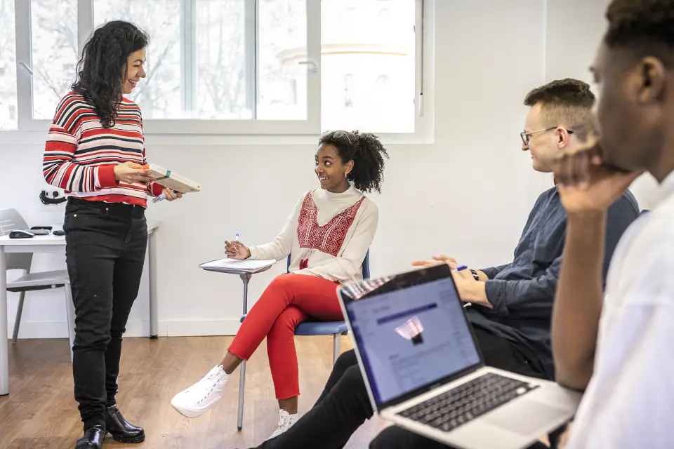 Three students sit at classroom desks while a professor leads a discussion.