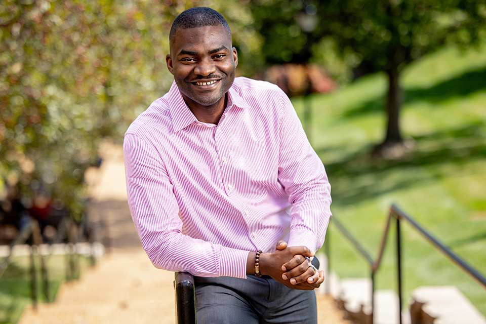 Dr. Oluwatoyosi Owoeye poses for a photo outdoors on a bright, sunny day. He wears a pink button-down shirt. He smiles at the camera. 