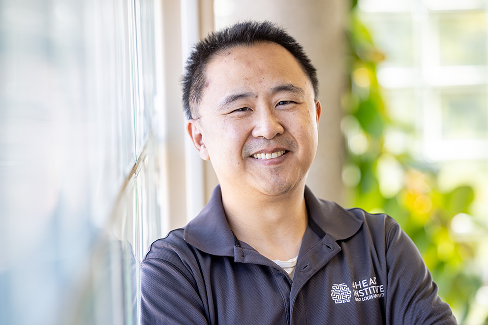 A man poses for a photo indoors next to a window. He smiles for the camera.