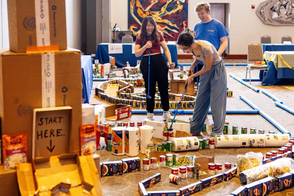 Two female and a male student play mini golf inside a room. The golf course is made out of cans and boxes of food.