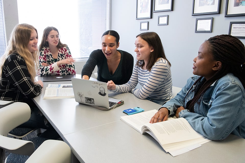 A study group of five women gather around a table in a room. An open lap top and books are on the table. 