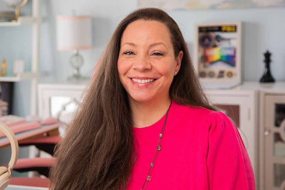 A head and shoulders picture of a woman facing the camera in a pink shirt with a living room seen in the background