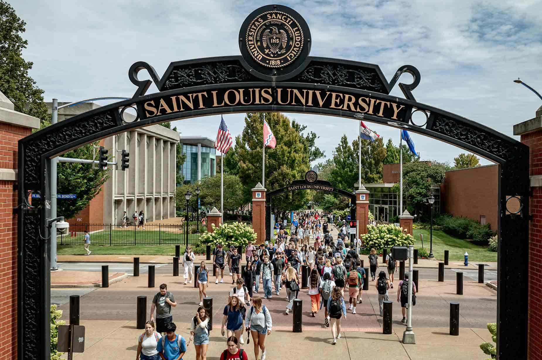 Students cross Grand Boulevard on a sunny afternoon. A pedestrian arch frames the view with “Saint Louis University” and the University seal visible at the top.