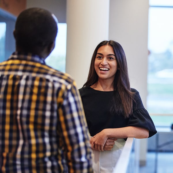 A student smiles and listens to another student speaking as they are standing up and talking next to a railing in the Saint Louis County Health Department.