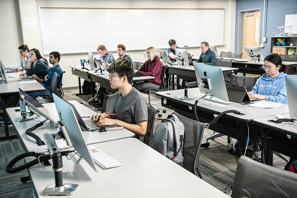 Students sit at desks in a classroom with laptops and computer monitors.