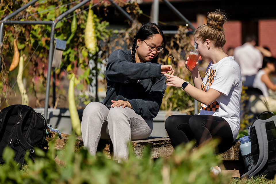 Two students sit on a bench outdoors. One student sips on a beverage while holding up a piece of food to share with her neighbor.