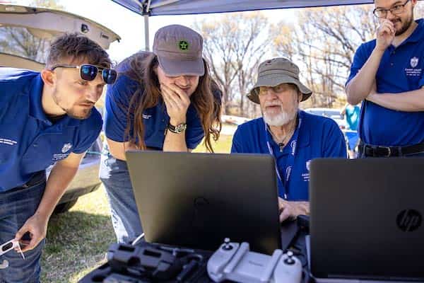 A male professor works on a laptop while three male students look on. The group is outside working under a tent and different equipment controllers can be seen in the foreground.