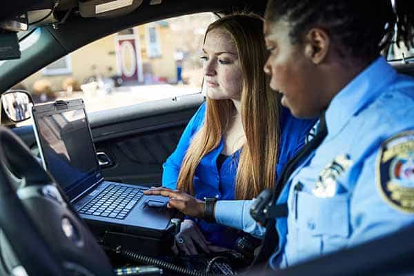 A female police officer works on a dashboard computer in a police squad care while a SLU student looks on.