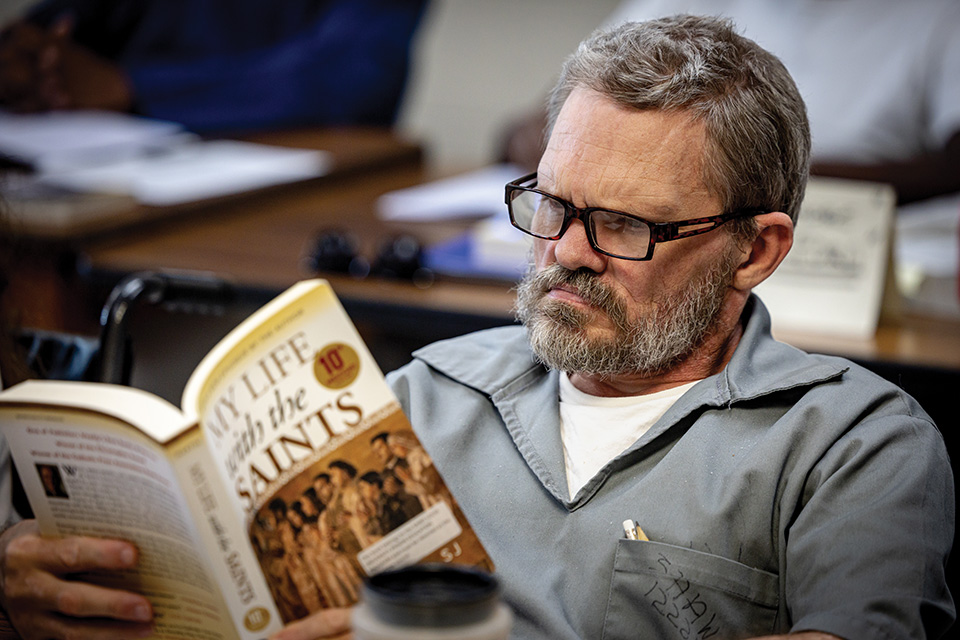 A white man with gray hair and a gray beard wears black-rimmed glasses and reads from My Life with the Saints