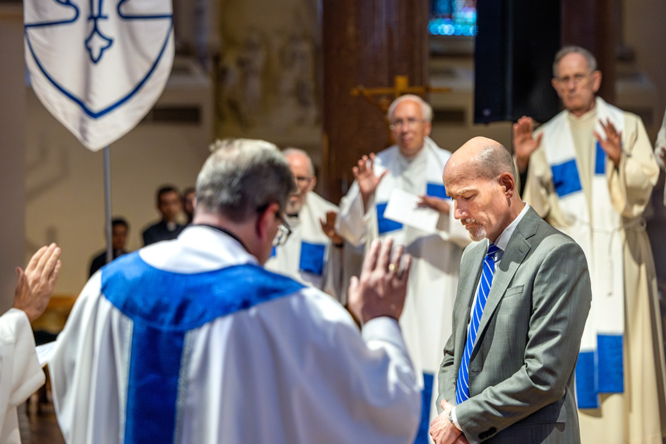 A bald white man in a grey suit stands in the middle of a group of Catholic priests in white garb.